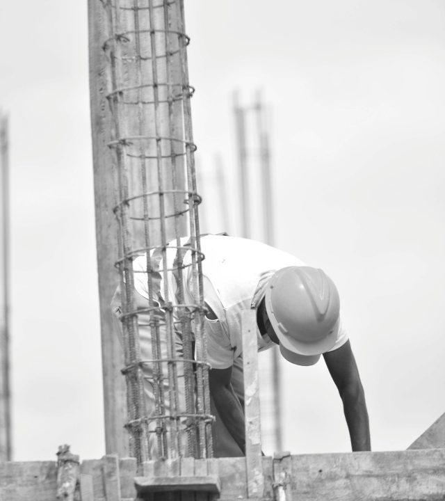 Grayscale image of a construction worker bending over a steel frame, taken outdoors.