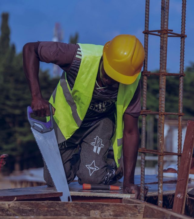 A construction worker in protective gear using a handsaw at a building site during the day.